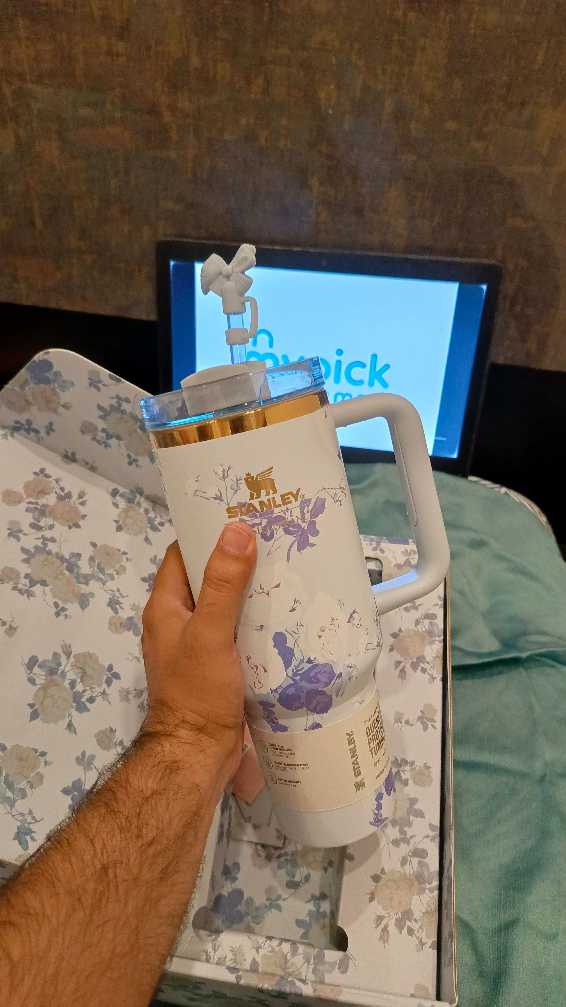 Hand holding a STANLEY mug with floral design in front of a television screen displaying a blue screen.