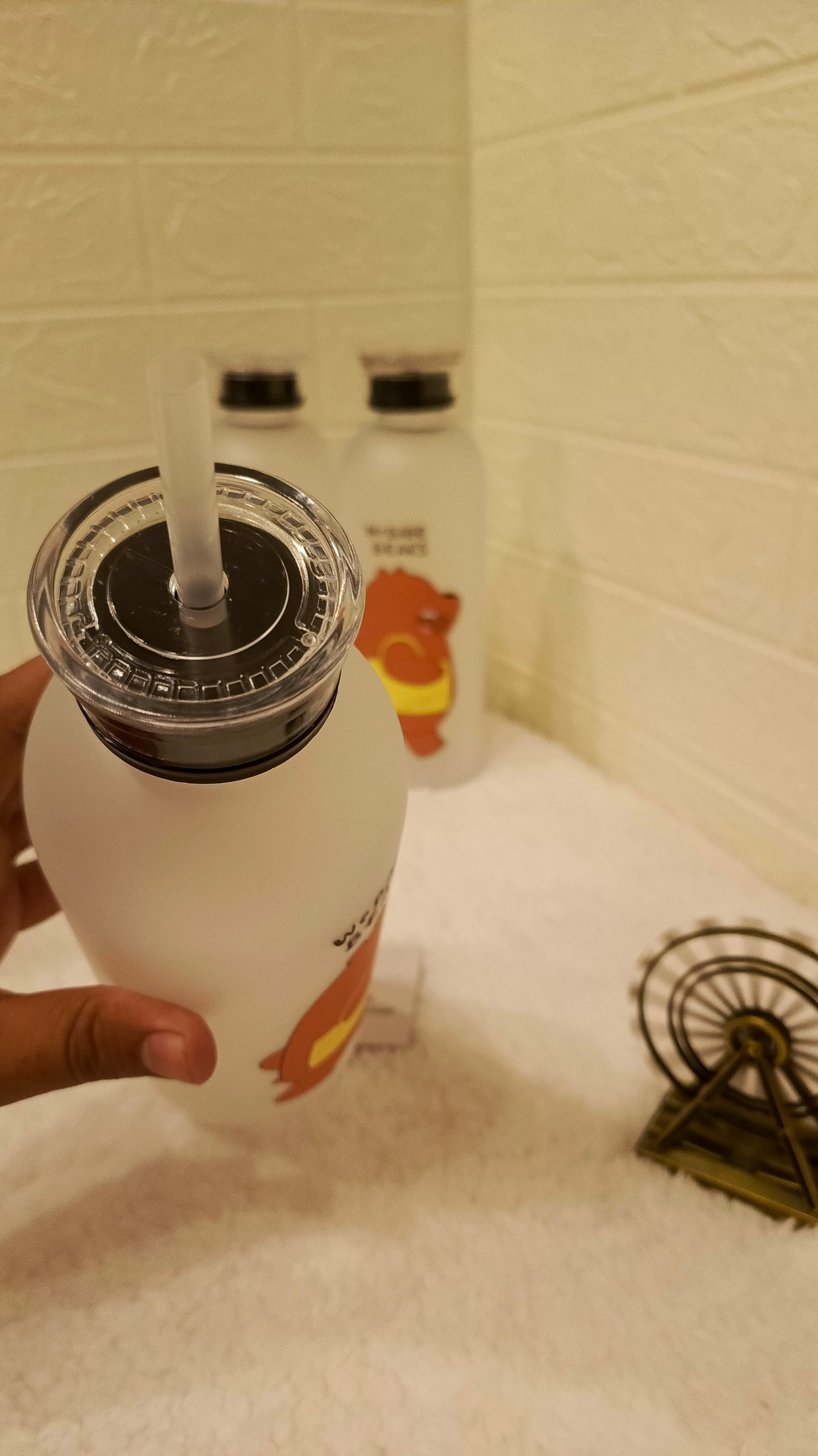 White bottle with black cap and straw held by a hand on a tiled bathroom counter.