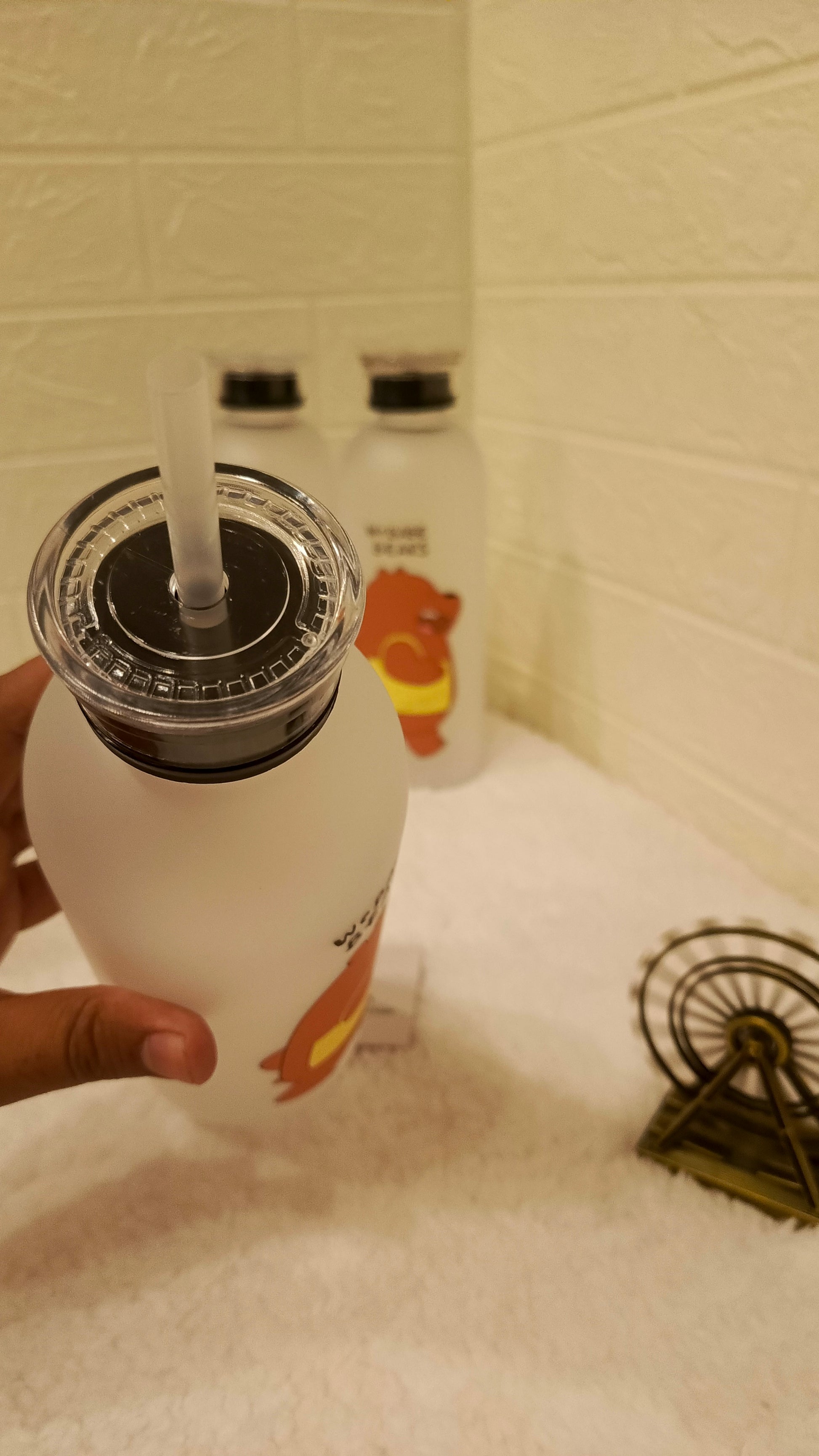 White bottle with black cap and straw held by a hand on a tiled bathroom counter.