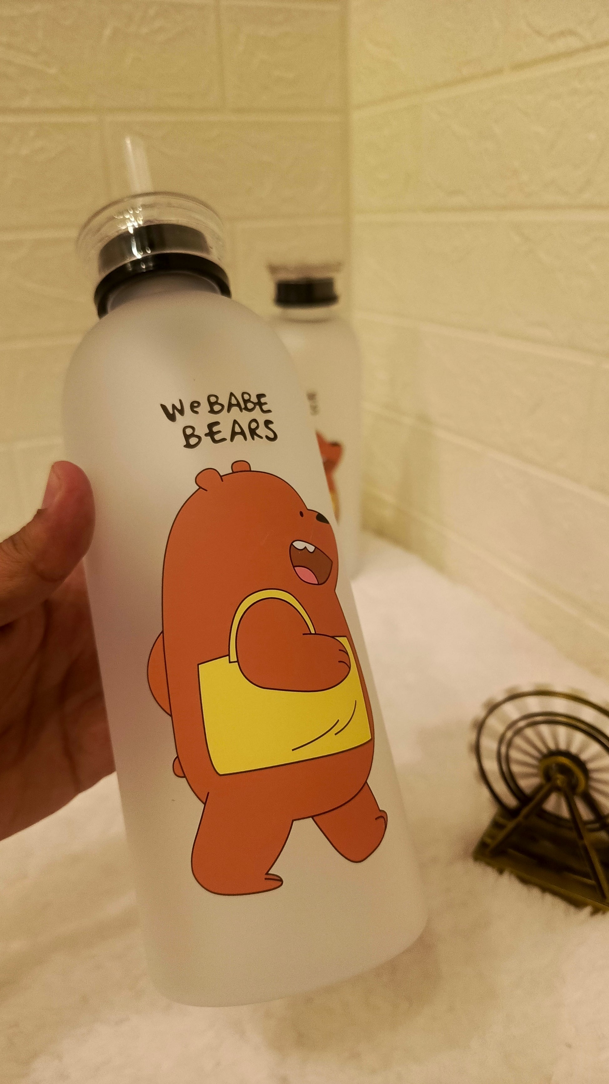 White water bottle with cartoon bear design and 'We Bare Bears' text, held against a tiled wall background.